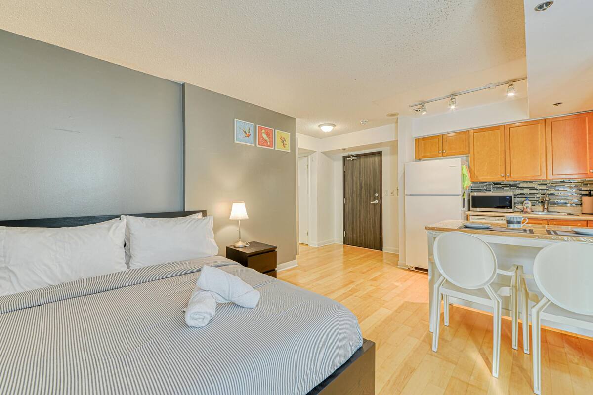 Habitación de hotel con cama, mesita de noche, lámpara y una zona de kitchenette con microondas, refrigerador y sillas.