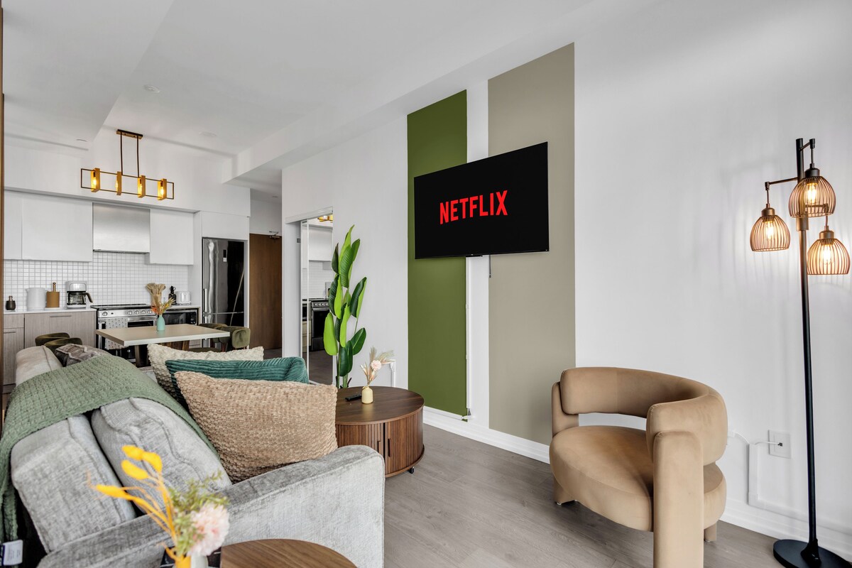 Uma sala de estar moderna com televisão de tela plana na parede, um sofá, uma mesa de centro e uma área de jantar ao fundo.