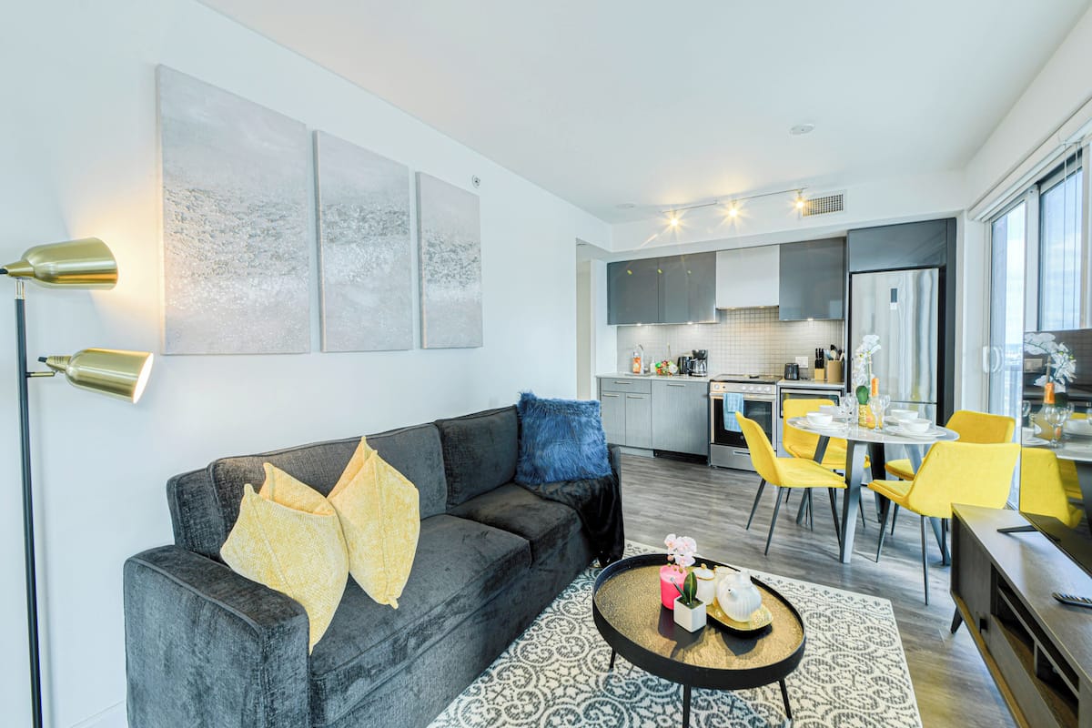 Una sala moderna con un sofá gris, sillas amarillas y una mesa de comedor. Se ve una cocina al fondo con gabinetes grises y una encimera blanca.