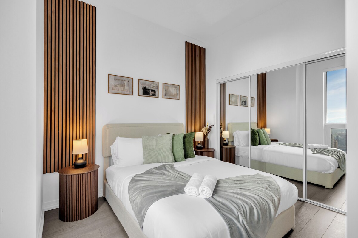 Quarto de hotel moderno com uma cama grande, mesa de cabeceira com abajur, espelho e obras de arte na parede.