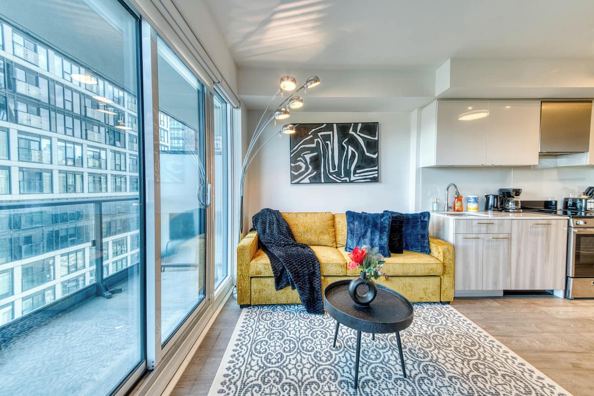 Una sala de estar moderna con un sofá, una mesa de centro y vistas a un paisaje urbano a través de amplios ventanales.