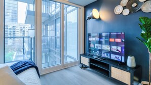 A modern bedroom with a large window, a flat-screen TV, a bedside table, and a decorative wall piece.