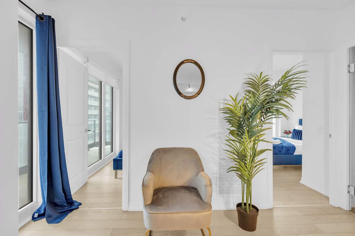 Una sala de estar moderna con un ventanal grande, una cortina azul, un sillón color beige, una planta en maceta y un espejo en la pared.