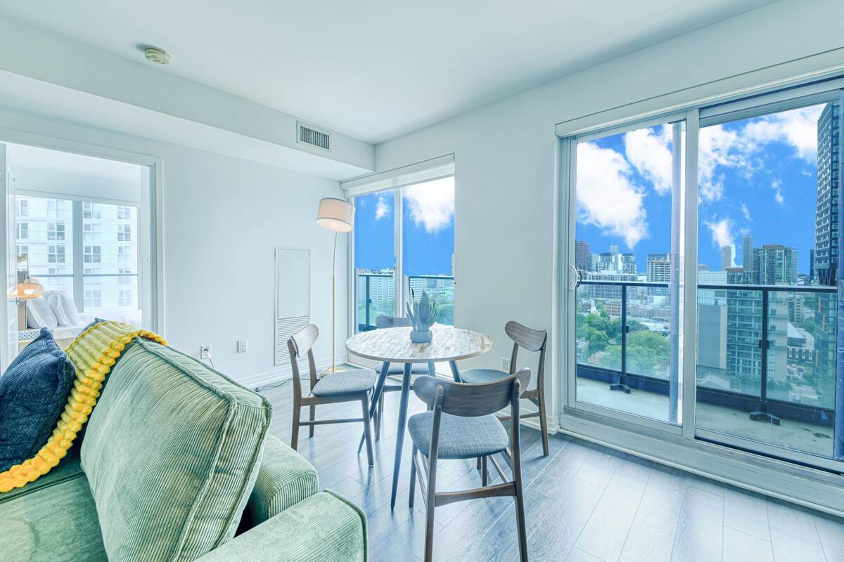 Una sala de estar moderna con un sofá verde, una mesa de comedor redonda con sillas y ventanas grandes que ofrecen vistas a la ciudad.