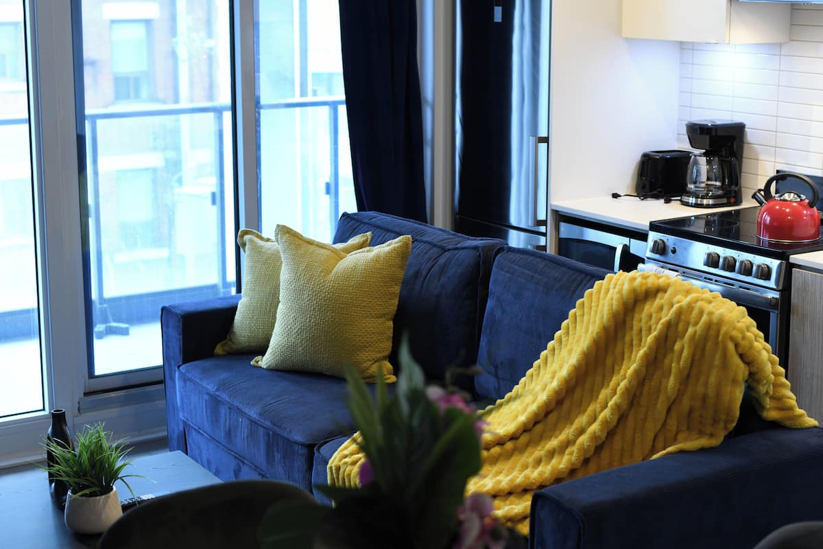 Un salón moderno con un sofá azul, un cojín amarillo y vistas de la ciudad a través de amplios ventanales.