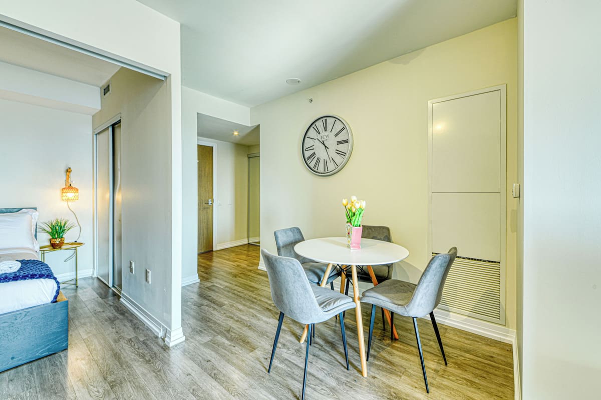 Una sala de estar moderna con comedor, que incluye una mesa redonda con cuatro sillas, un reloj en la pared y una cama con cabecera azul.