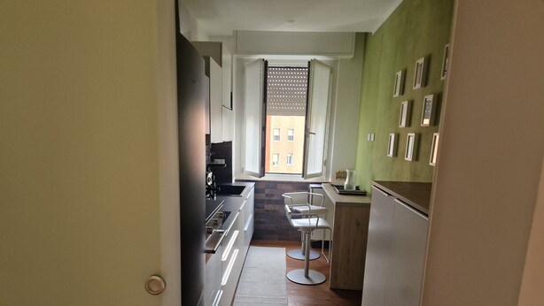 A narrow kitchen with a dining area, wooden flooring, and a view through a door to an outdoor balcony. - The houses of Vanda (Milano)