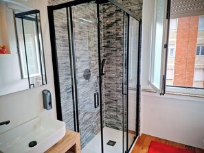 Ein modernes Badezimmer mit Duschkabine aus Glas, einem weißen Waschbecken und einem Fenster mit Blick auf ein Backsteinhaus.