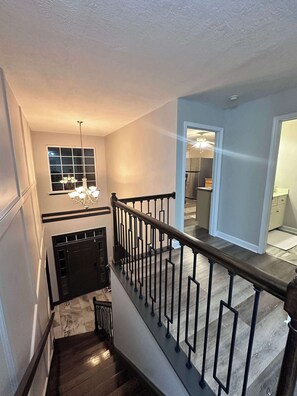 A staircase with a black railing, a chandelier, and a window with blinds. - Premier Executive Living | 4br Luxury Apartment (Evans)