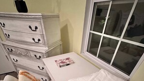 Habitació amb escriptori blanc, cadira amb coixí decoratiu, planta en test i finestra amb persianes.