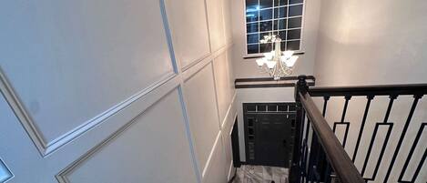 A staircase with a dark wooden handrail, white wall paneling, and a chandelier.