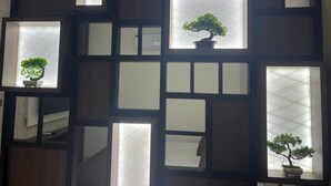 A room with a geometric wall design, multiple windows, and potted bonsai trees. - Nandha Grande (Udumalaipettai)