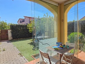 A glass-enclosed patio with a table and chairs, a hanging chair, and a view of a green lawn and buildings in the background. - Two-room apartment Limonaia in Marina di Campo (Marina di Campo)