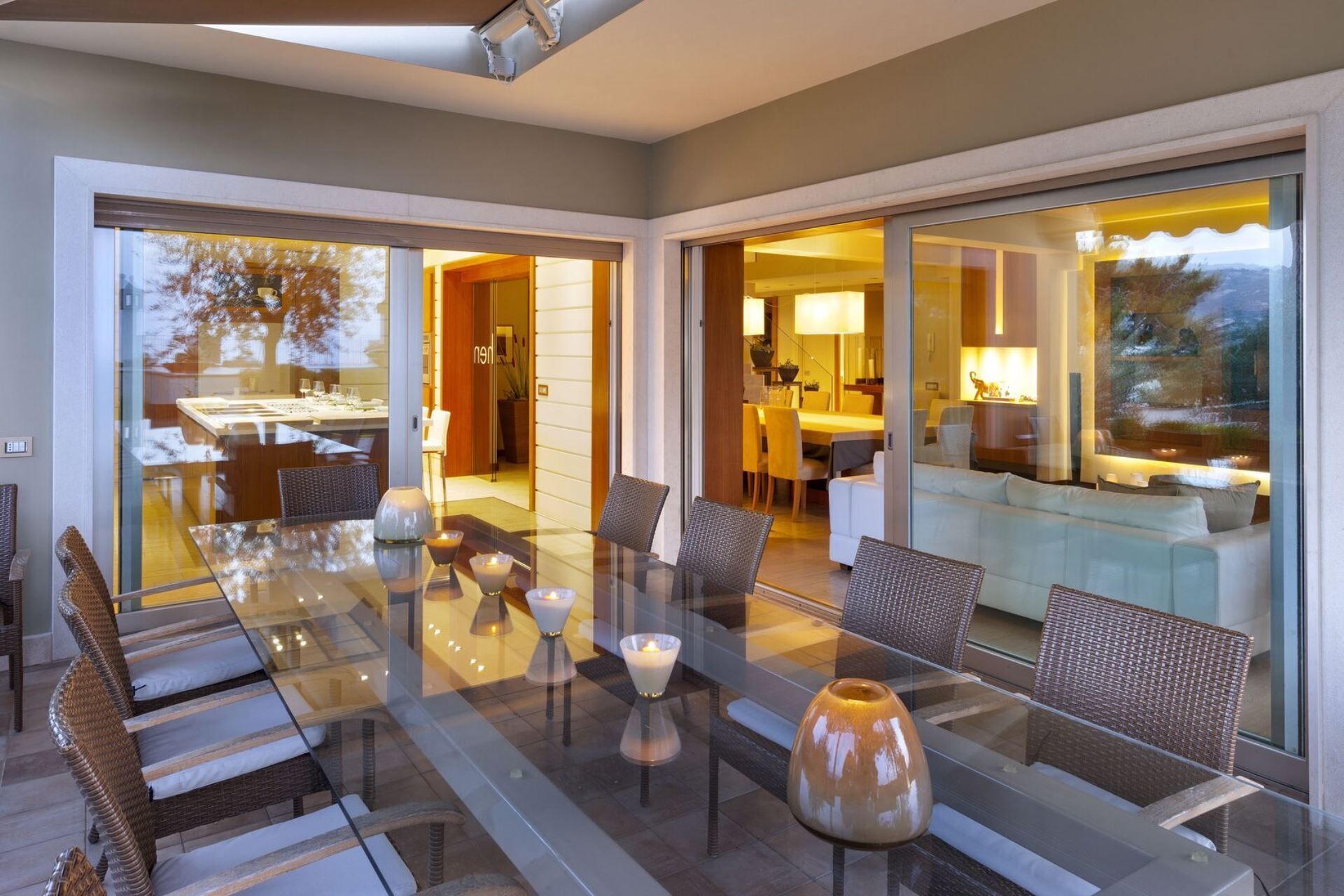 A modern dining area with glass tables and wicker chairs, a glass-enclosed kitchen, and a living area with a sofa and lamp.
