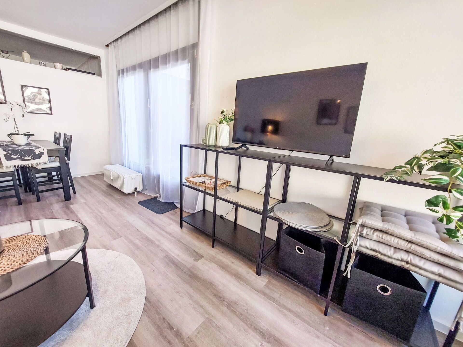 A modern living room with a flat-screen TV, a dining area with a table and chairs, and a large potted plant.