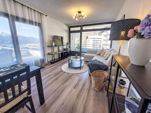 A modern living room with a sofa, a TV, a dining table, and a large window offering a view of the city. - Lovely apartment for 6 people with WIFI, TV and balcony (Bad Gastein)