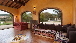 A room with a red sofa, a patterned sofa, and a red armchair. There are two large windows with arched tops, a wooden ceiling, and a tiled floor. - Umbria Genius Loci Farmhouse (Collazzone)