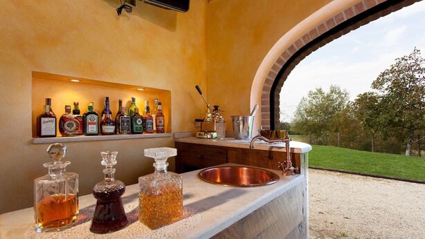 A bar area with a brick oven, a shelf with various liquor bottles, and a copper sink. - Umbria Genius Loci Farmhouse (Collazzone)