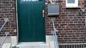 Una puerta verde con un ventanal encima y una ventana pequeña a la derecha, y una escalera de hormigón que sube hacia la puerta.