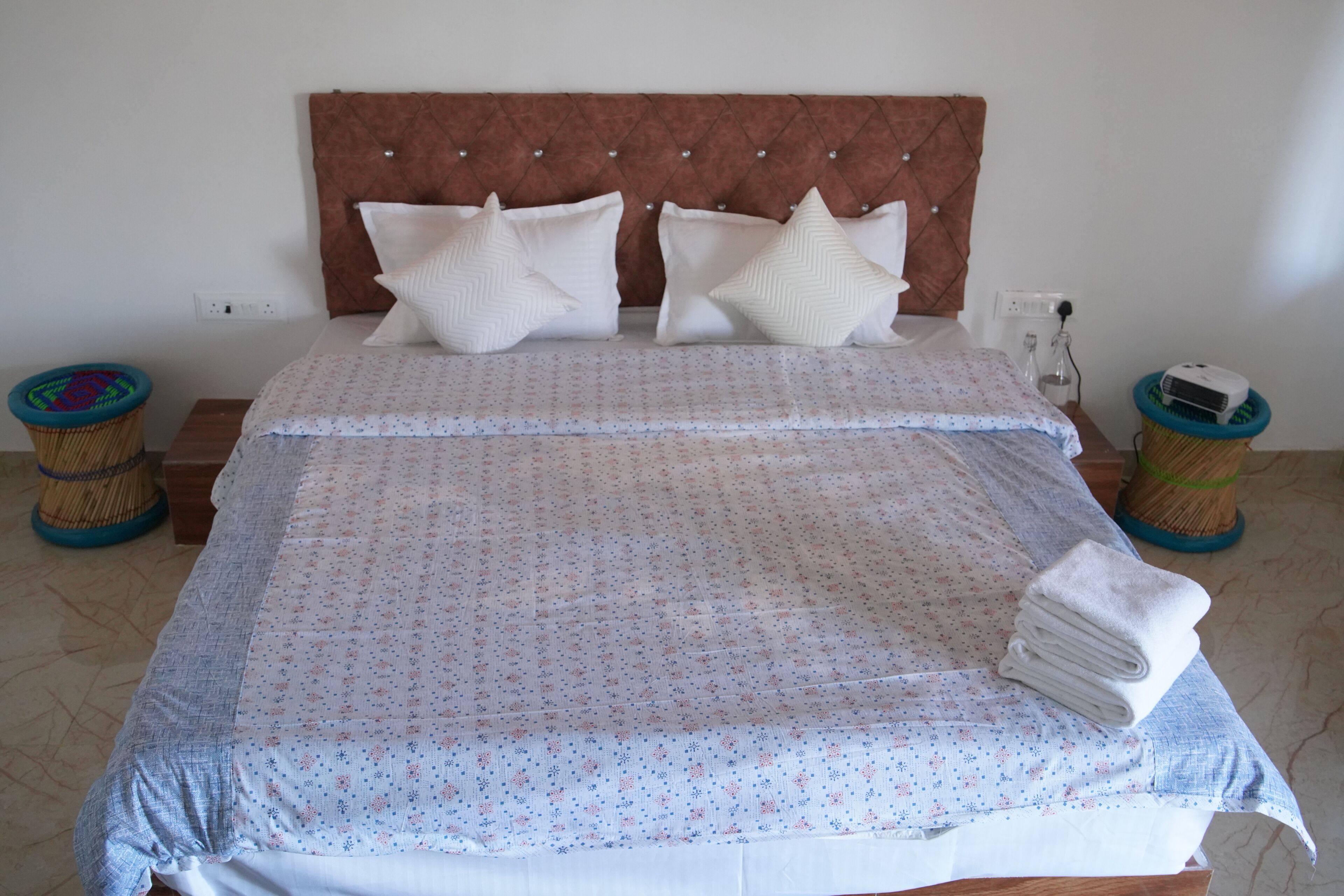 A neatly made bed with a patterned bedspread, two decorative pillows, and a stack of folded towels on a wooden nightstand.
