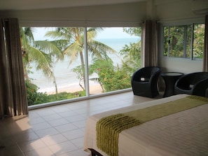 Una habitación con una cama, dos sillas, una mesita y un ventanal grande con vistas a una playa y palmeras.