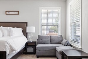 A bedroom with a bed, bedside table, lamp, grey sofa, and a window with blinds.