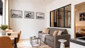 A modern living room with a grey sofa, a dining table with chairs, a coffee table, and a large window with a view of a building. - TheBlueStay 350 - Stunning Apt in République (Paris)
