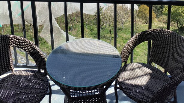 Un balcón con sillas de mimbre y una mesa de vidrio con vistas a un paisaje montañoso que incluye casas y campos verdes.