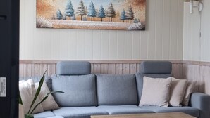 A living room with a grey sofa, a wooden coffee table, a painting of trees, and a lamp on the wall. - Cozy 2-bedroom cabin in peaceful Gryllefjord with AC (GRYLLEFJORD)