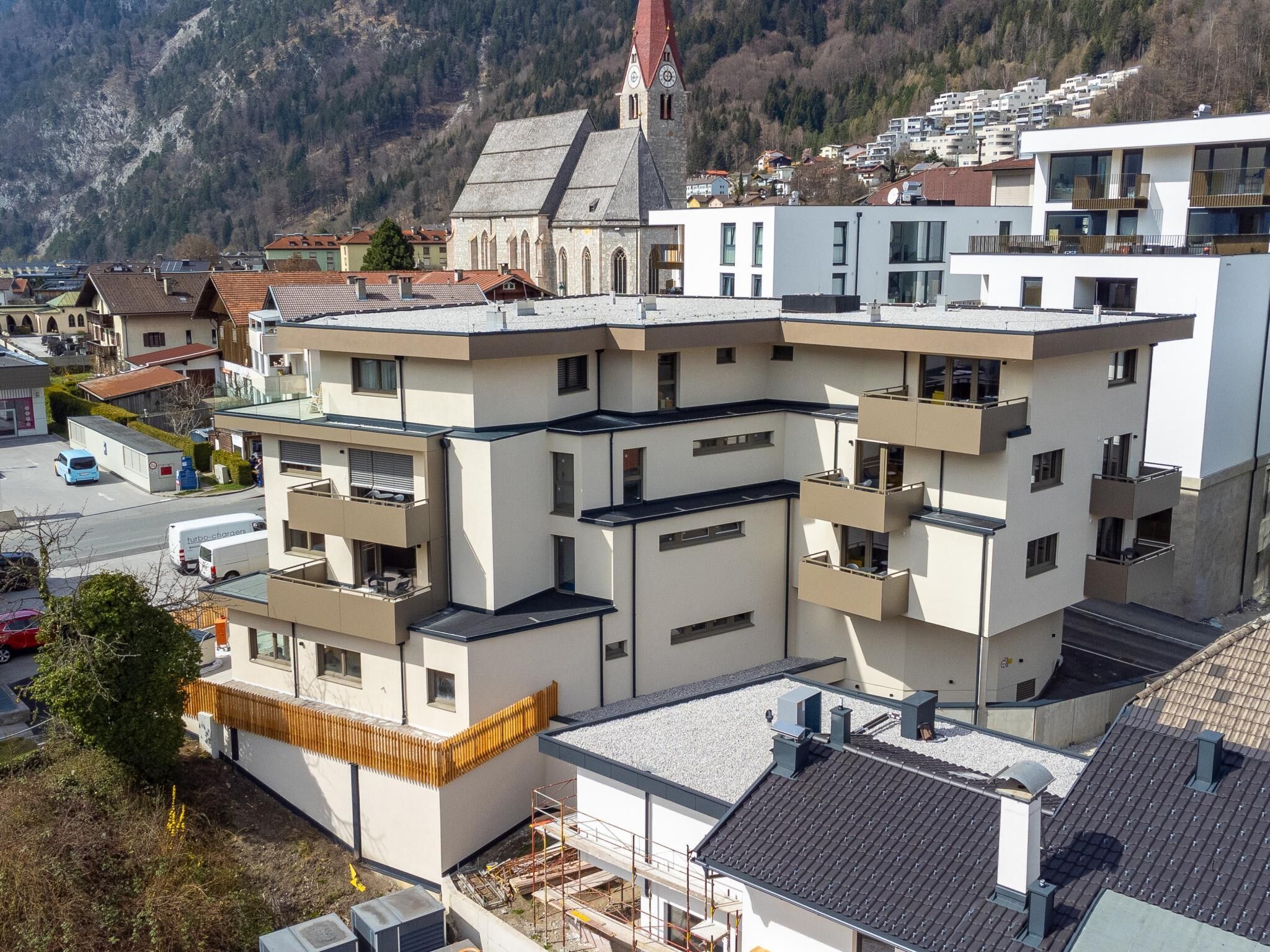 Ein mehrstöckiges Gebäude mit Balkonen, eine Kirche mit einem Glockenturm und eine gebirgige Kulisse.