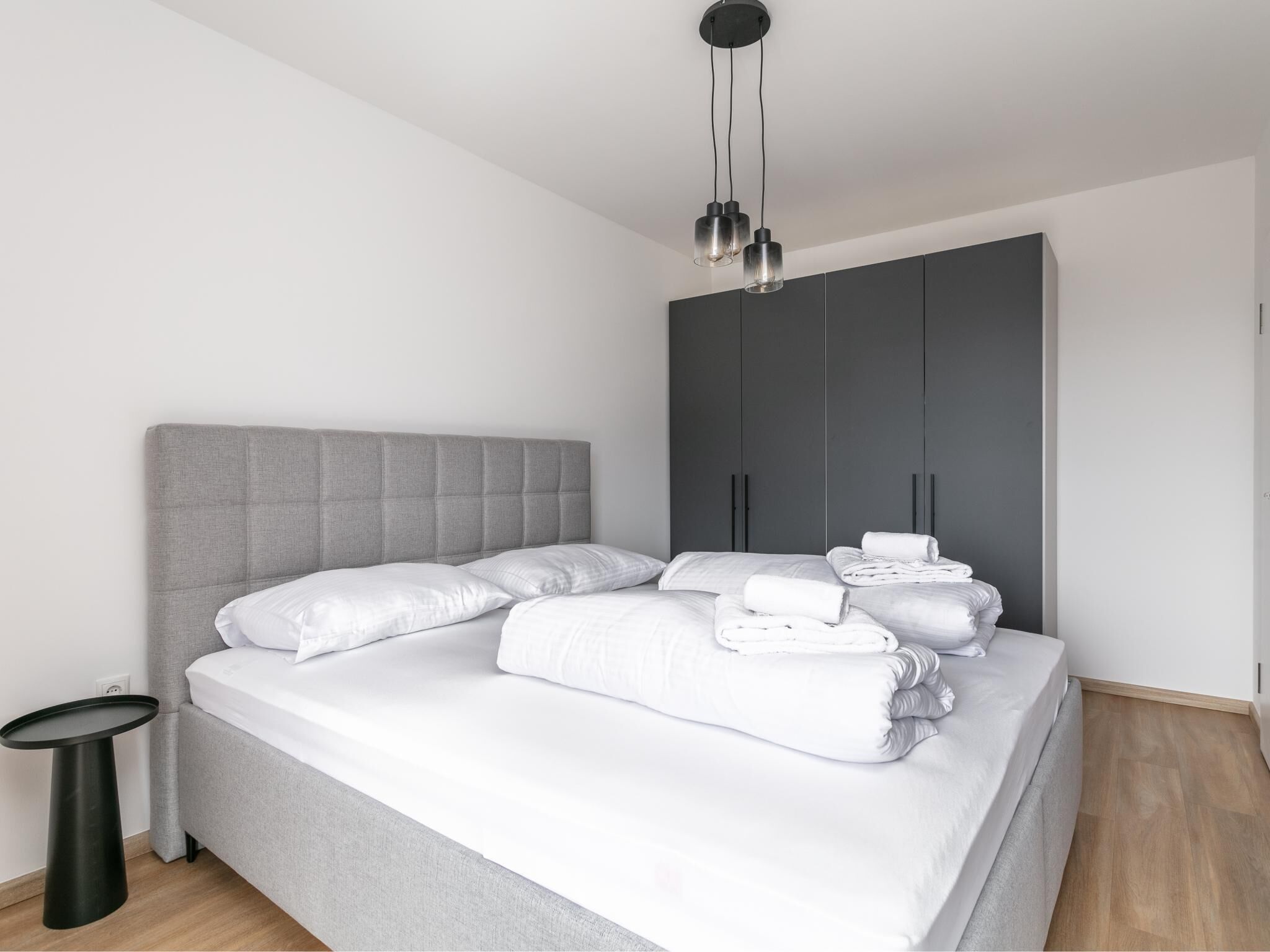 Ein modernes Schlafzimmer mit grauem Stoff-Kopfteil, einem Bett mit weißen Bettbezügen, einem grauen Kleiderschrank und einem schwarzen Nachttisch.