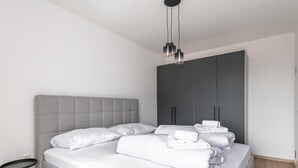 Ein modernes Schlafzimmer mit grauem Stoff-Kopfteil, einem Bett mit weißen Bettbezügen, einem grauen Kleiderschrank und einem schwarzen Nachttisch.