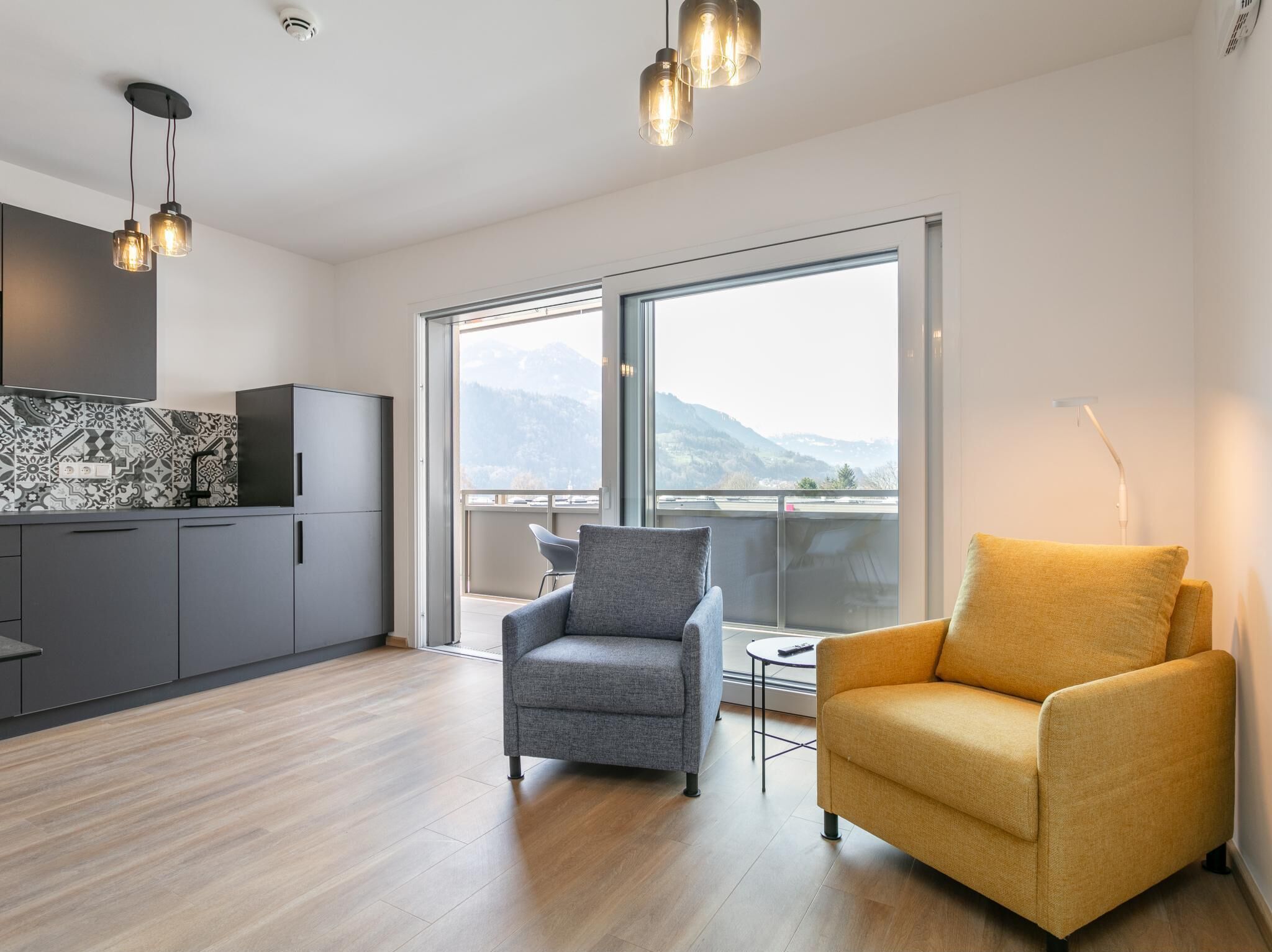 Ein modernes Wohnzimmer mit grauem und gelbem Sessel, einer Küche mit dunklen Schränken und einem großen Fenster mit Blick auf die Berge.