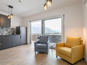 Ein modernes Wohnzimmer mit grauem und gelbem Sessel, einer Küche mit dunklen Schränken und einem großen Fenster mit Blick auf die Berge.