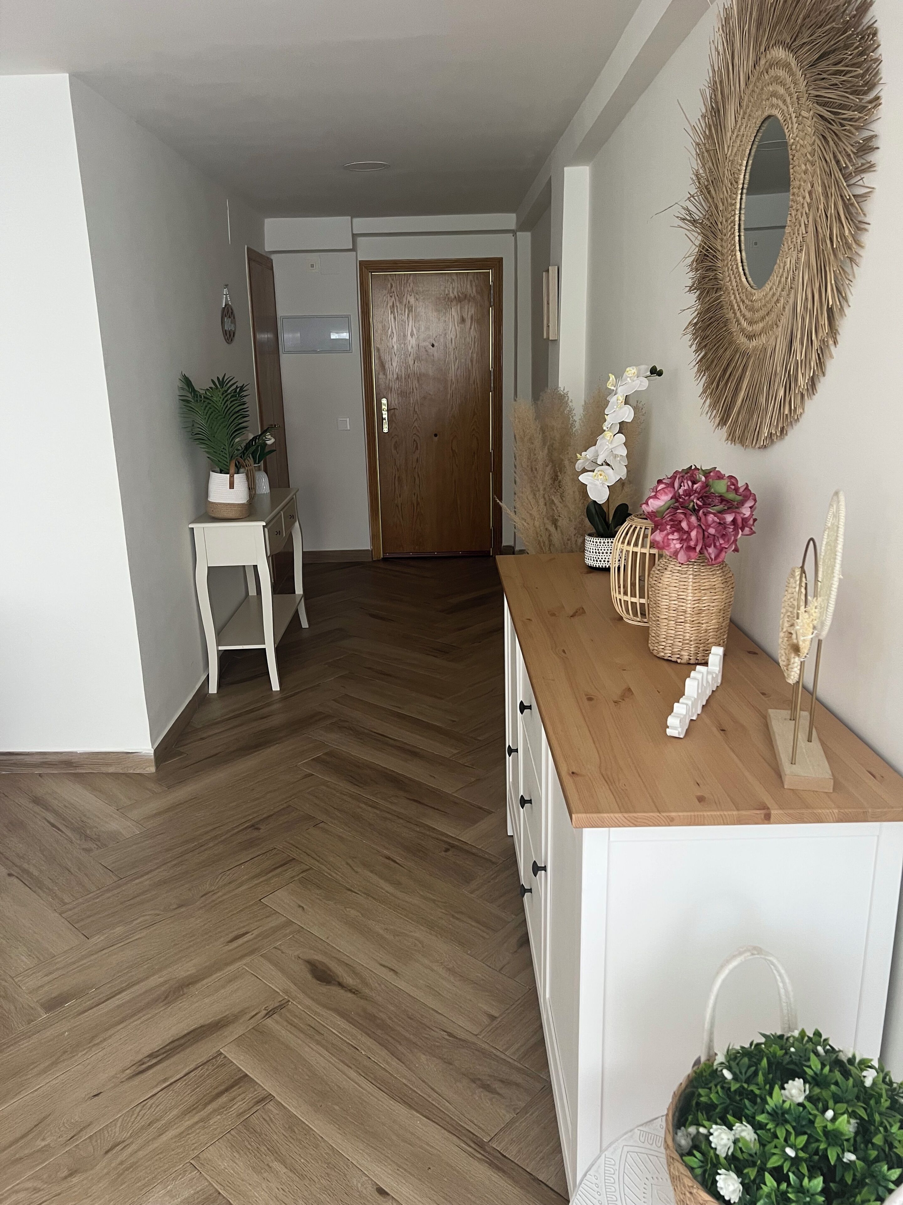 Un couloir avec un plancher en bois, une armoire blanche avec des tiroirs, une petite table avec une plante, et une porte au fond.