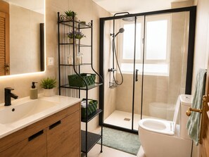 A modern bathroom with a glass-enclosed shower, a white toilet, a wooden vanity, and a shelf with plants and storage containers. - Peaceful 3-bedroom apartment in Salou near the beach (Salou)