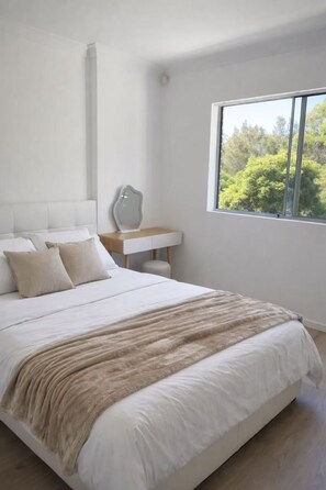 Een slaapkamer met een groot bed, een nachtkastje, een spiegel en een raam met uitzicht op bomen.