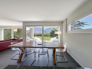 Een eetgedeelte met een houten tafel en stoelen, een rode bank en uitzicht op bergen door grote ramen.