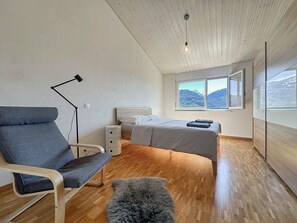 Een slaapkamer met een bed, een stoel, een lamp, een kleedje en een raam met uitzicht op bergen.