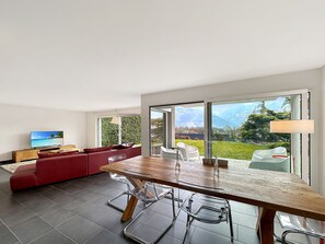 Ein modernes Wohnzimmer mit einem großen Esstisch aus Holz, transparenten Glasstühlen, einem roten Sofa und einem Fernseher auf einem Holzgestell.