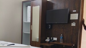 A hotel room with a bed, a TV mounted on a wooden panel wall, a small desk with a phone and a water bottle, and a trash bin.