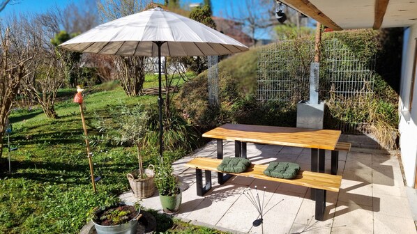 Eine Terrasse im Freien mit einem Holztisch und einer Bank, umgeben von viel Grün und einem Metallzaun.