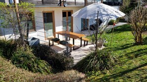 Une terrasse avec une table en bois et des bancs, entourée de verdure et agrémentée d’un parasol blanc.