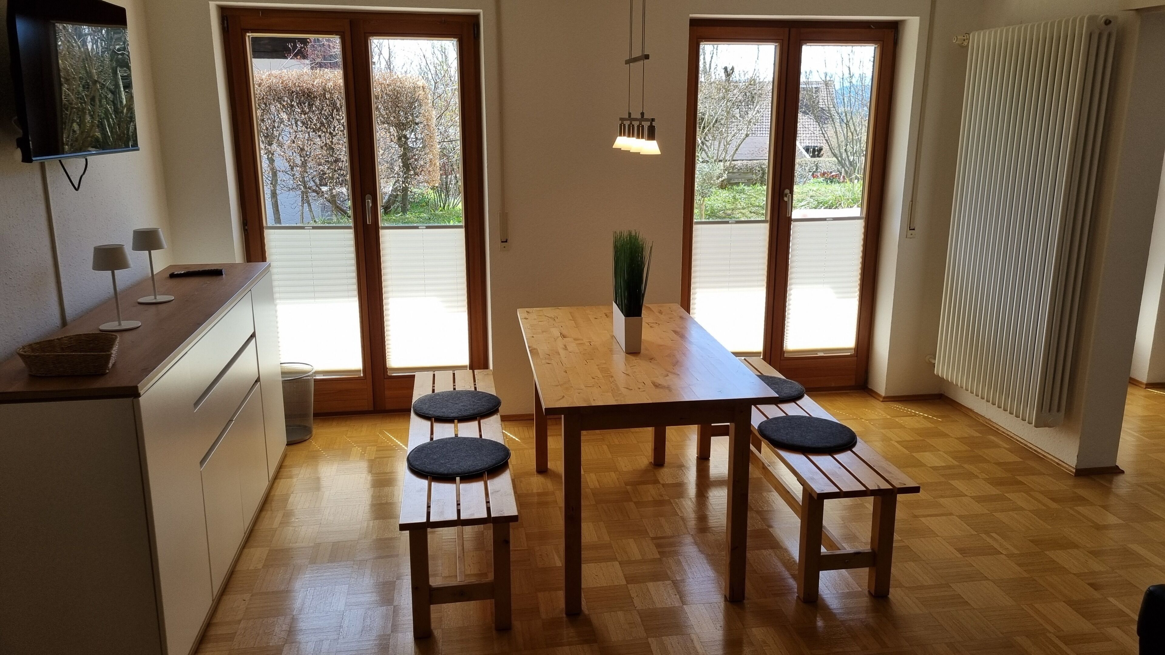 A modern kitchen with a wooden table and benches, a TV mounted on the wall, and large sliding glass doors leading to a garden.