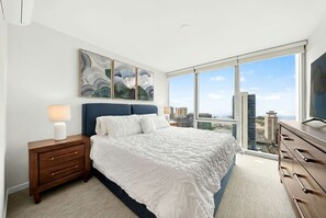 Ein modernes Hotelzimmer mit einem großen Bett, einem Nachttisch, einer Kommode und Blick auf die Stadt durch große Fenster.