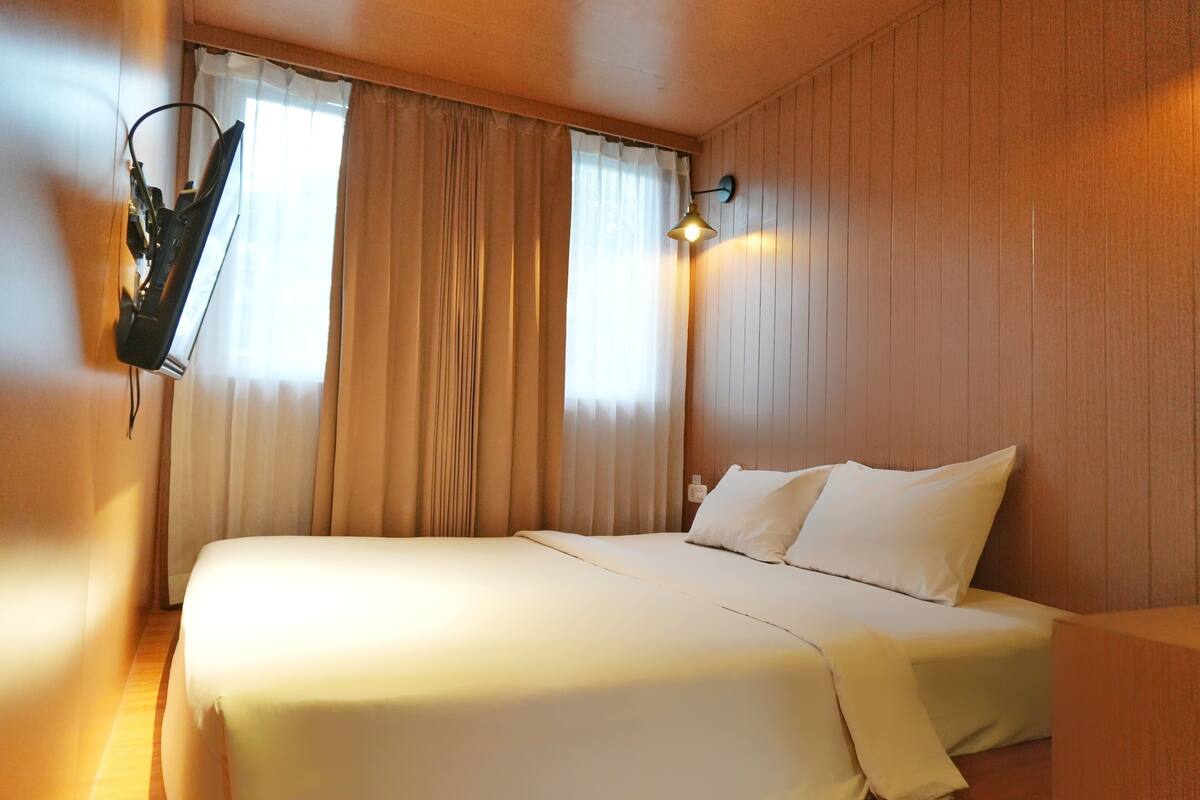 Una habitación de hotel con cama, cortinas, una lámpara fijada a la pared y un panel de madera.