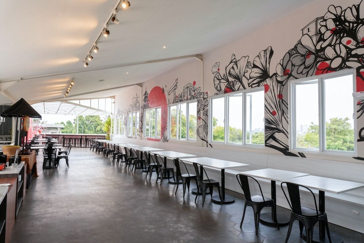 Un espacio interior amplio con mesas y sillas largas, ventanas grandes y un mural en la pared.