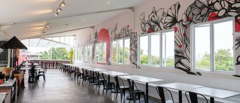 Un espacio interior amplio con mesas y sillas largas, ventanas grandes y un mural en la pared.