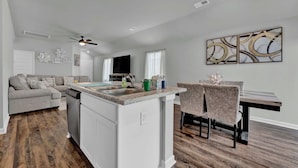 A modern kitchen with a central island, a dining area with a table and chairs, a sofa, and a ceiling fan.