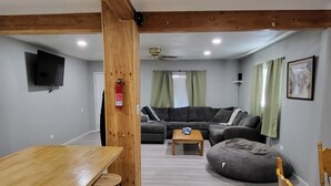 Una habitació amb una taula de dinar de fusta, dos tamborets d'alta, un sofà, un seient de bossa i una taula de centre de fusta petita, i una televisió penjada a la paret.
